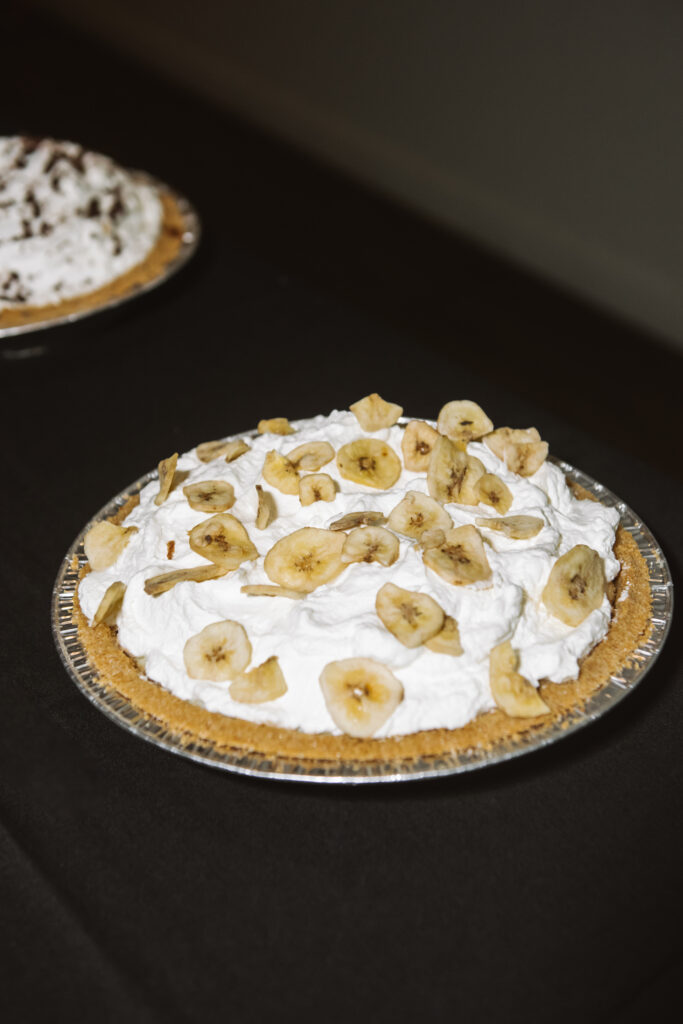 Close up of a banana cream pie on a black tablecloth. There is an unidentifiable mousse/cream pie in the background on the same table.