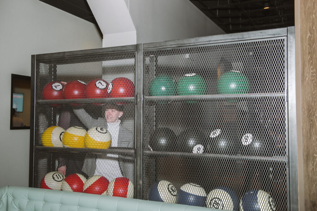 Bowling ball case featuring multiple bowling balls of different colors and numbers. There is one person seen through the metal case about to select a bowling ball.