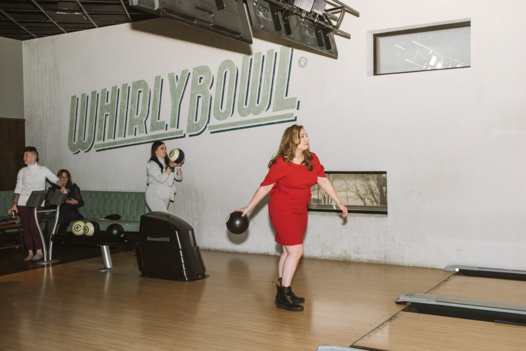Bride seen pre-bowling ball throw. The bowling ball is in her right hand, being held behind her while her left hand is counter-balancing that action. "Whirlybowl" is painted on the wall behind her and there are wedding guests seated/standing behind the bride and one has a bowling ball in her hands.
