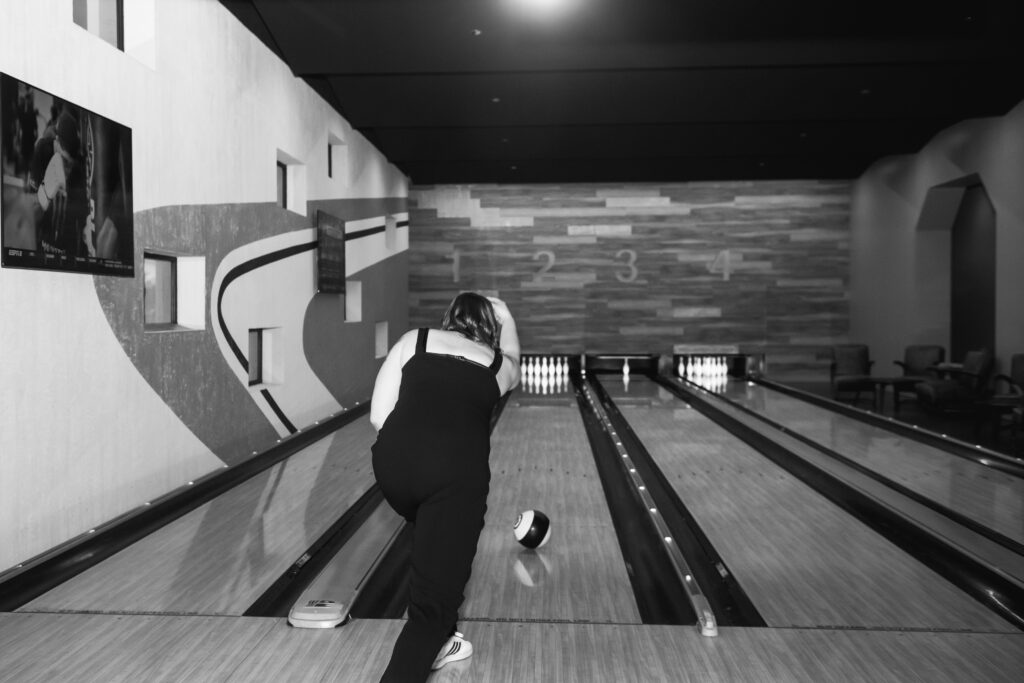 Black and white photo of a bride shown from behind having just thrown a bowling ball that is rolling in the lane. Her right hand is still outstretched from throwing the ball and she is leaning forward towards the lane.