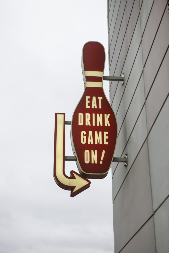 Whirlyball exterior sign in the shape of a bowling pinball with the words "EAT DRINK GAME ON!" written on it. There is an arrow to the sign's left that points down and right.