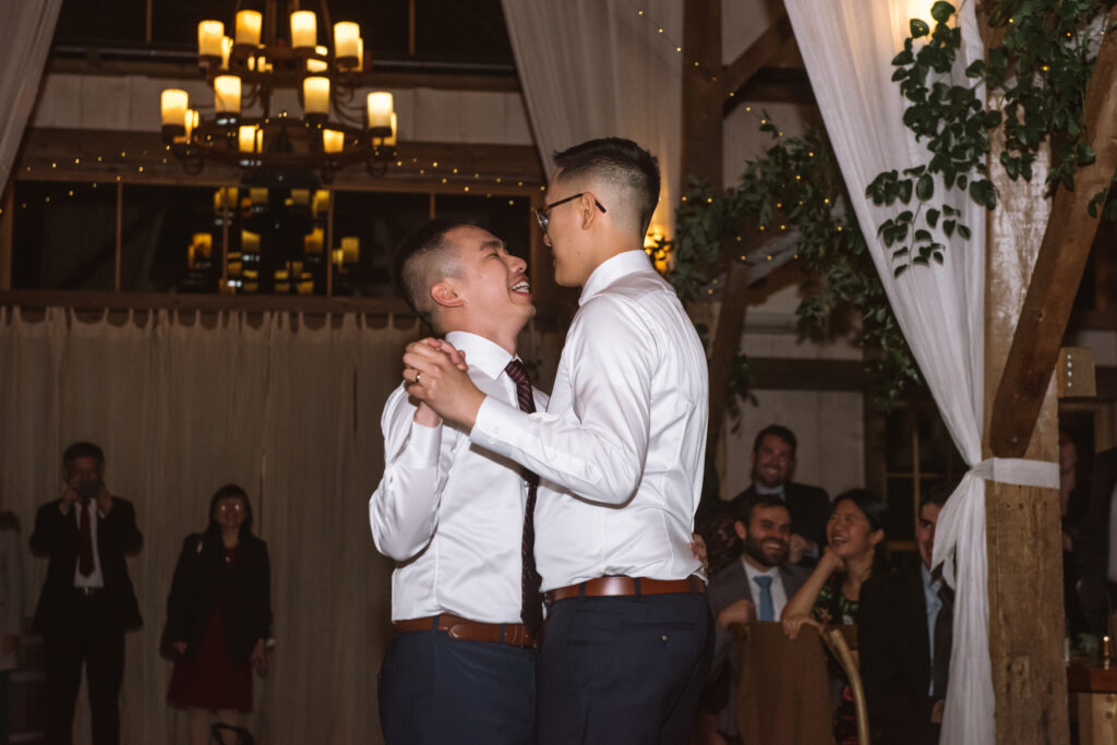 Two grooms slow dancing during their first dance as husbands during their reception. They are standing facing one another and are holding and embracing each other. They are both smiling, looking at one another.