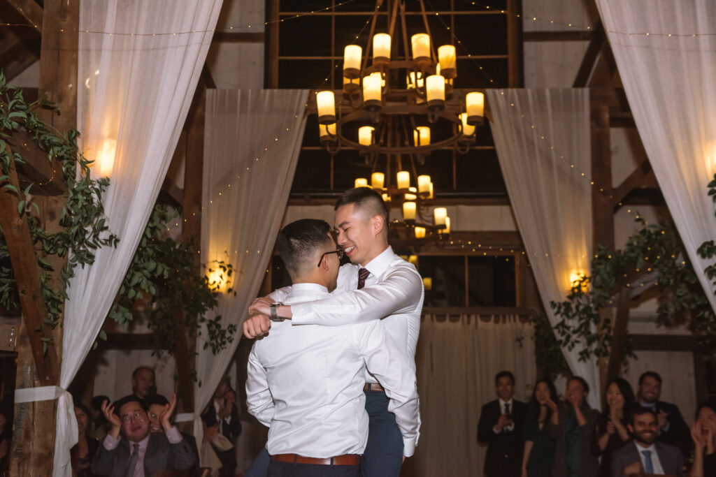 Two grooms dancing during their first dance at their reception. One is holding the other who has his legs wrapped around his new husband's waist. The one being carried is smiling down at his husband and has his arms around his shoulders. Guests are seen clapping and smiling in the background.