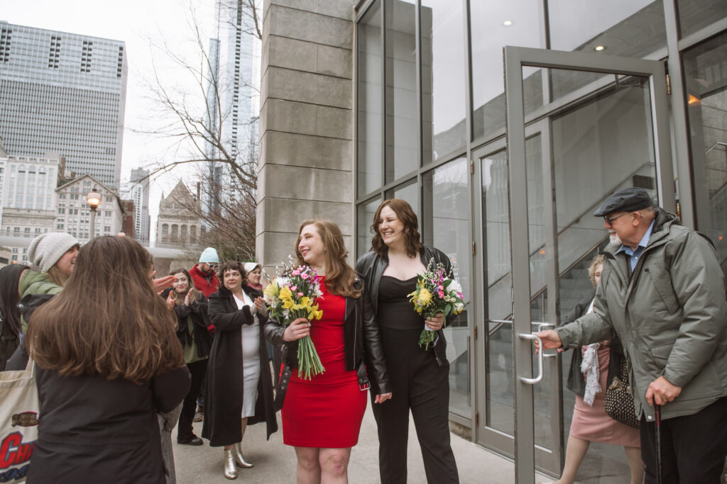 Two brides walking side by side walking out of a building being greeted by friends and family. Both are holding their bouquets in hand and are smiling and looking to their right.