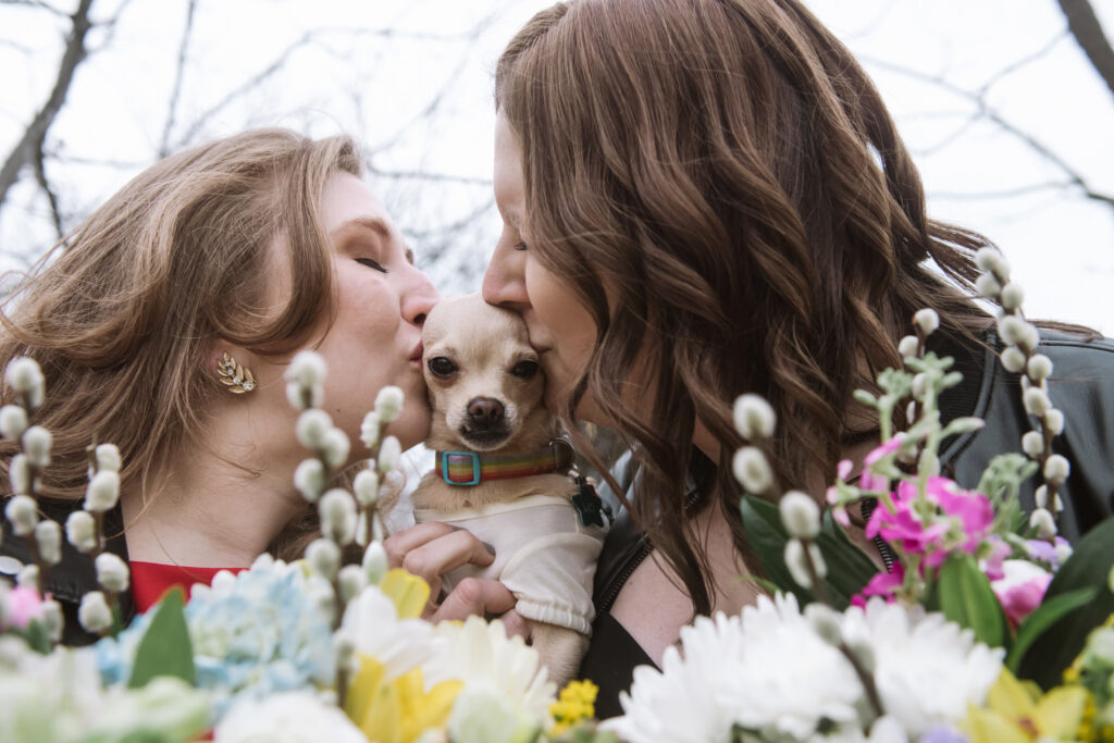 Close up of two brides holding their little Chihuahua in between them. Their Chihuahua is facing the camera and her lower right canine is visible in her closed mouth. She is dressed in a shirt with a rainbow colored collar. Both brides are giving their dog a kiss, eyes closed, from either side of the dog. Their bouquets are visible in the bottom foreground with trees/tree branches in the background.