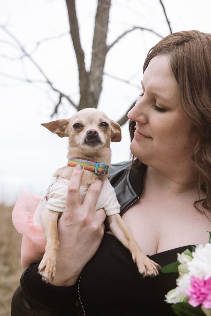 Bride holding her little Chihuahua up near her face. The bride is looking at her dog with a soft smile and the dog is looking slightly off camera in a slightly disgruntled fashion. Her eyes are turned more backwards and her lower right canine is visible through her closed mouth.