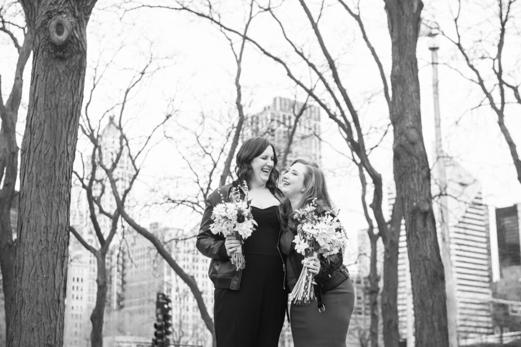 Black and white photo of two bride standing side by side. They are smiling wide-open-mouthed and holding a bouquet each. There are trees surrounding them and Chicago buildings in the background.