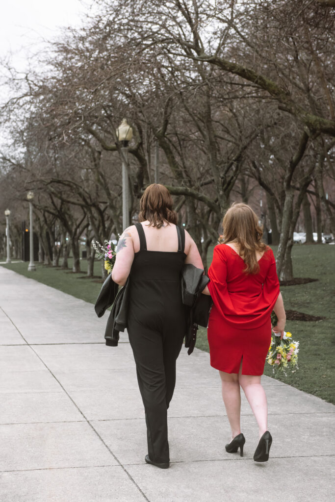Two brides seen from behind. One has her left arm wrapped in the other's right arm. They are walking away from the camera.