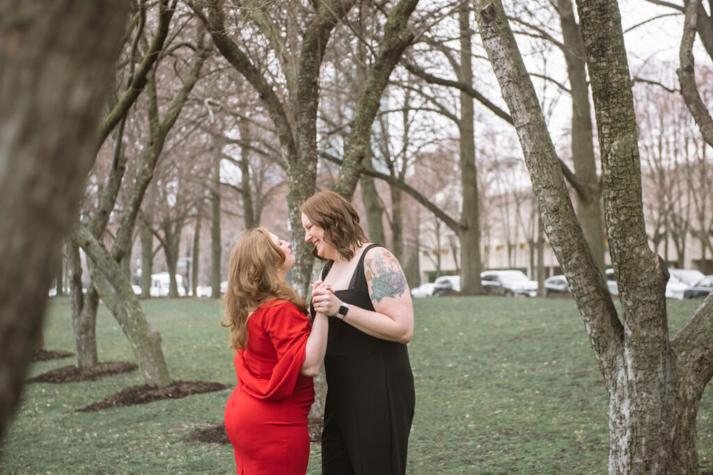 Two brides standing facing one another mid-slow dance. They are smiling at one another and are surrounded by trees in a park.
