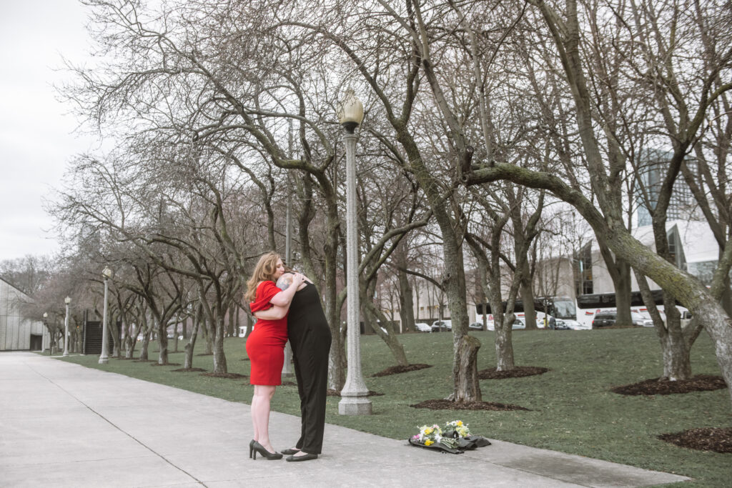 Two brides hugging each other, facing one another standing on a park walkway with trees in the background.