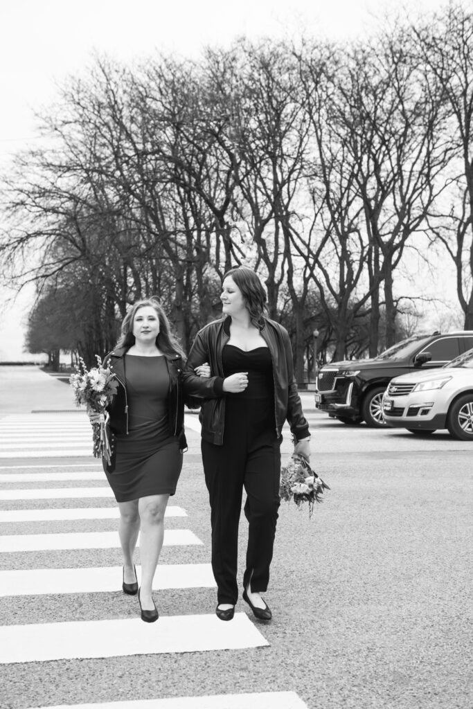 Black and white photo of two brides, arm in arm, walking on a city crosswalk. There are cars and trees in the background. One is facing the camera and the other is softly smiling at her wife.