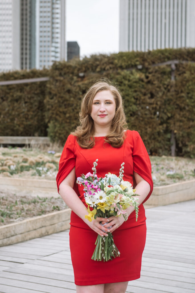 Close up of a bride wearing a sleek and modern red dress with an attached cape/wrap holding her bouquet in front of her. She is standing in a park with hedges in the middle ground and Chicago buildings in the background. She has a soft closed-mouth smile and is directly looking to the camera.