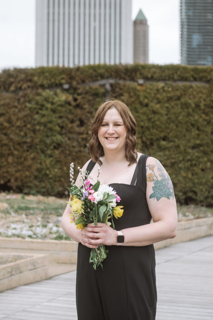 Close up torso portrait of one of the brides in a sweet-heart neckline black jumpsuit holding her bouquet in front of her. She is smiling directly to the camera. She is standing in a garden with a hedge in the middle ground and Chicago buildings in the background.