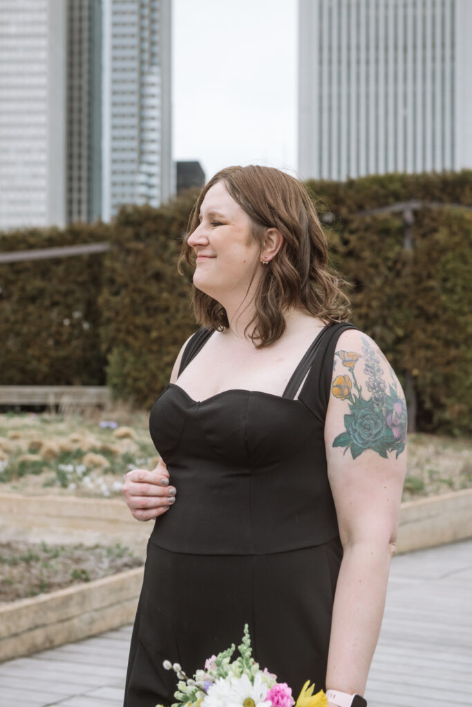 Close up torso portrait of one of the brides in a sweet-heart neckline black jumpsuit holding her bouquet loosely on her left side. Her right hand is sitting lightly on her waist. She is smiling softly off camera to her right. She is standing in a garden with a hedge in the middle ground and Chicago buildings in the background.