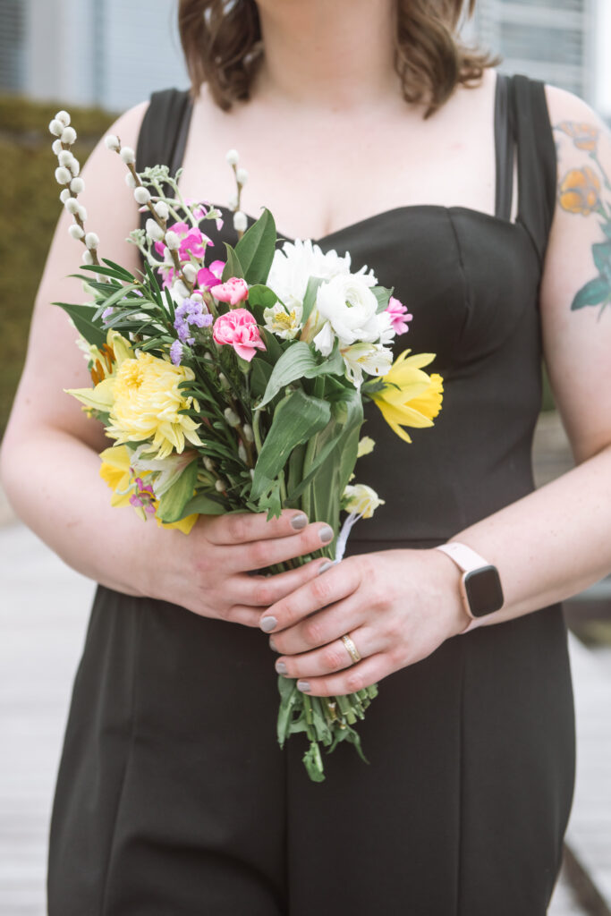 Close up of a bride wearing a black sweetheart-neckline black jumpsuit holding her bouquet in front of her. Only her torso and upper legs are framed in the photo to showcase the bouquet.