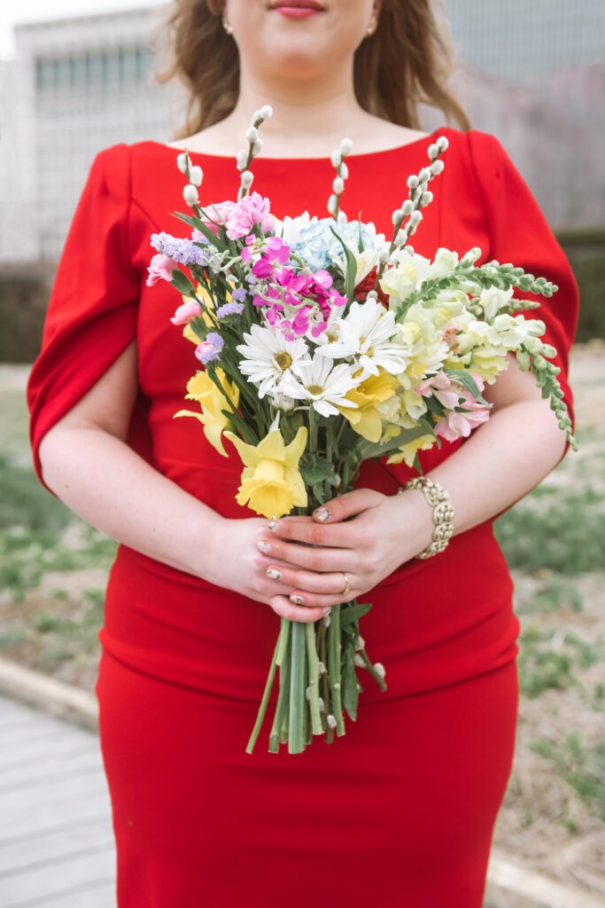 Close up of a bride wearing a sleek and modern red dress with an attached cape/wrap holding her bouquet in front of her. Only her torso and upper legs are framed in the photo to showcase the bouquet.