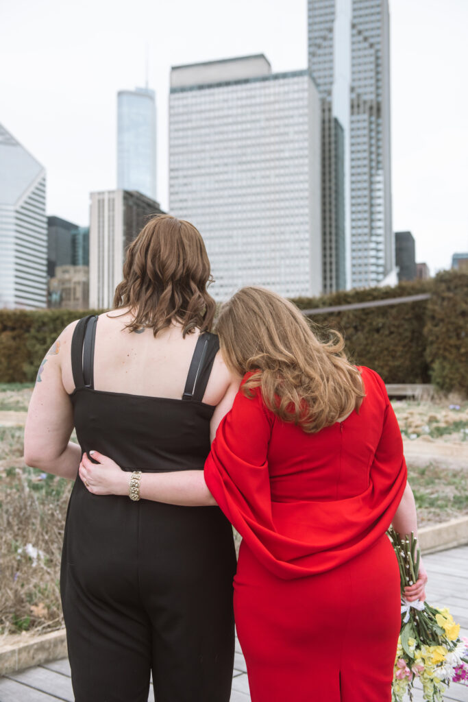Two brides standing side by side with one having her arm across the other's back and resting her head on the other's right shoulder. They are facing away from the camera viewing the Chicago skyscrapers in the background.