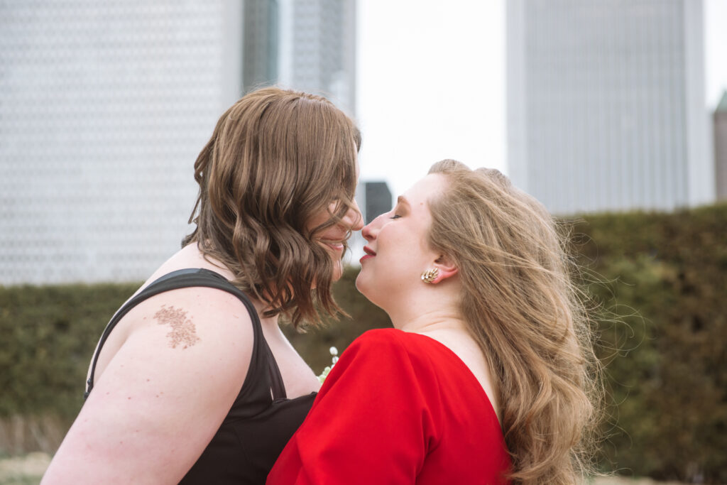 Portrait of two brides embracing one another facing each other. They are in an almost-kiss. Their eyes are closed and their mouths are slightly open in a soft smile. They are standing in a park with hedges in the middle ground and Chicago buildings in the background.