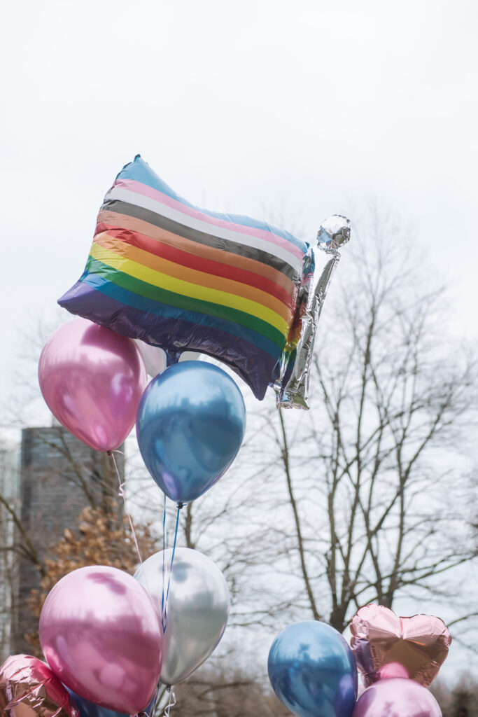 Close up of pride balloons. One is in the shape of a flag and it is the pride flag which incorporates the black and brown stripes as well as the trans flag. The other balloons are blue, pink, and white in reference to the trans flag colors. There are traditional shaped balloons as well as pink heart-shaped balloons.