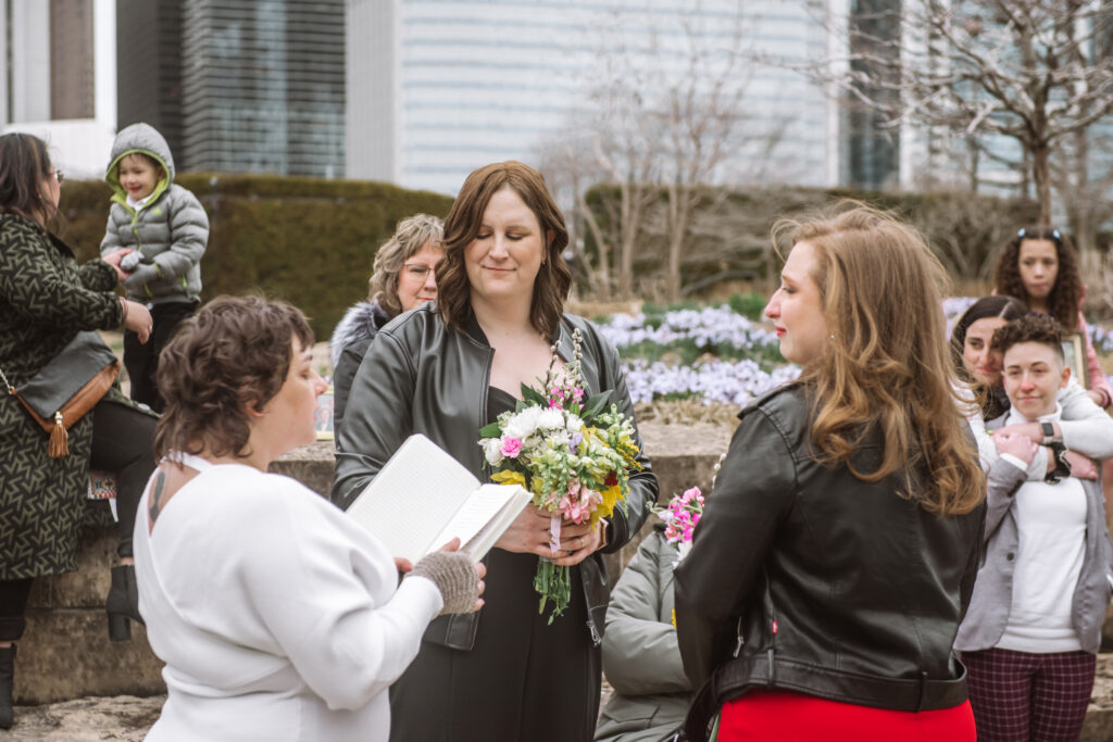 Two brides and their officiant mid-ceremony. The officiant is reading from an open journal. There are guests surrounding them and city buildings in the background.