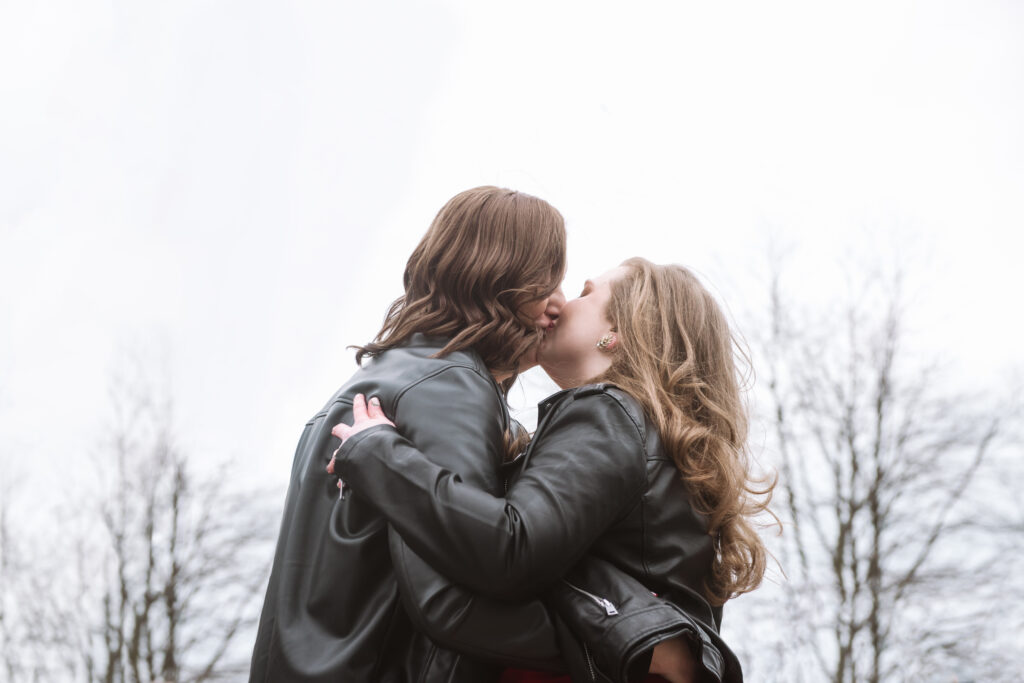 Two brides kissing after being pronounced wives. They have their arms wrapped around one another in a passionate embrace. There are trees in the far background.