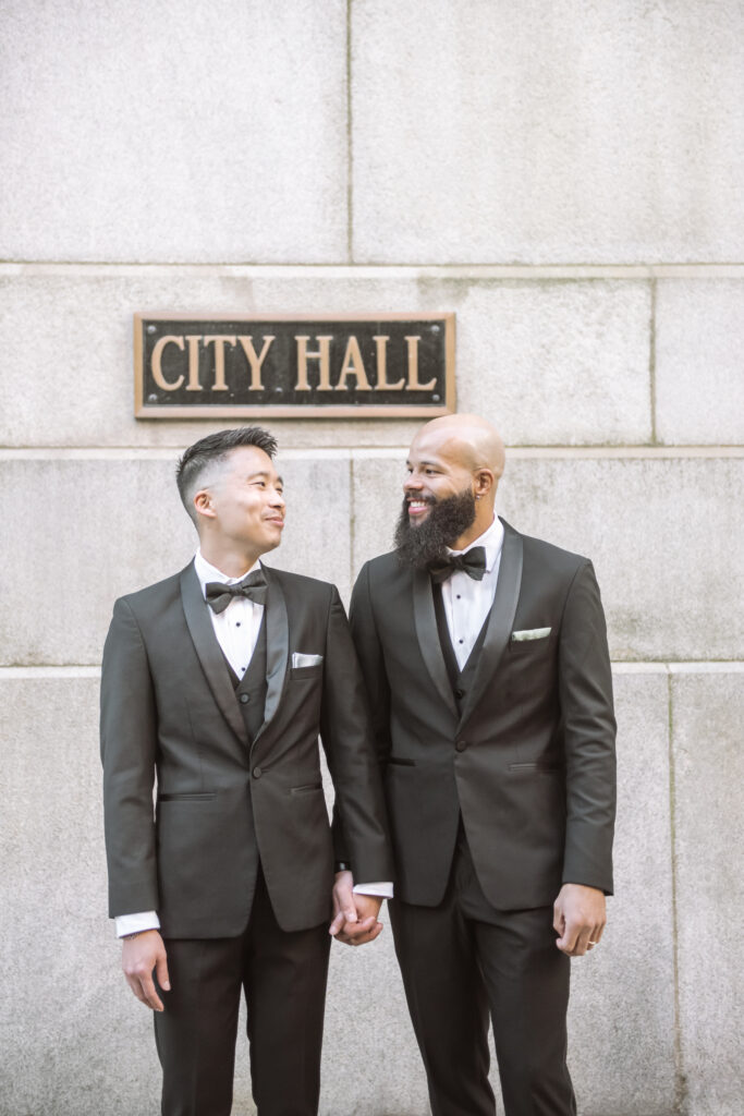 Two marriers, both in black tuxes, standing side-by-side holding hands in front of the Chicago City Hall sign.