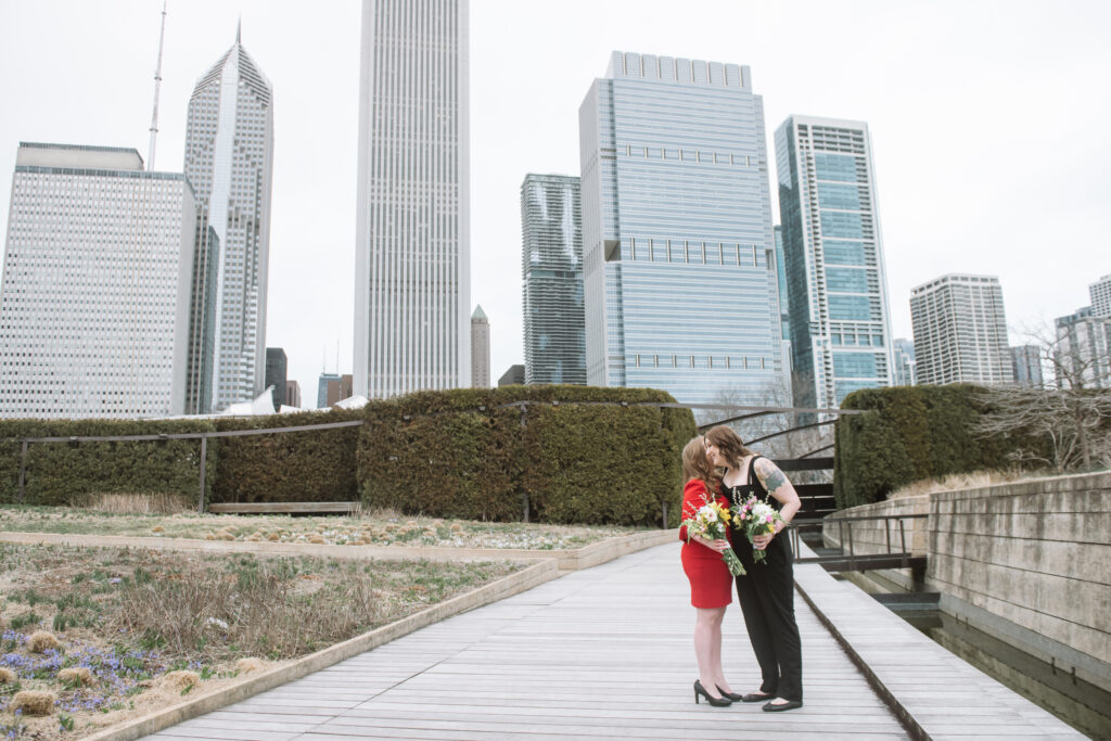 Britt, she/her, wearing a red dress & holding a bouquet talks into the ear of Rya, she/her, wearing a black jumpsuit & holding a bouquet in front of Chicago skyline