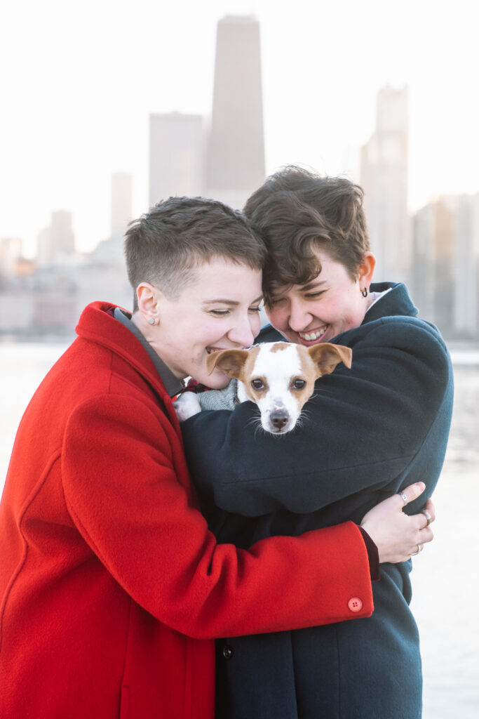 A couple standing facing one another, one with their arms around the other who is holding their small terrier mixed breed rescue dog. They are both smiling and looking at their dog who is looking to the camera with floppy ears.