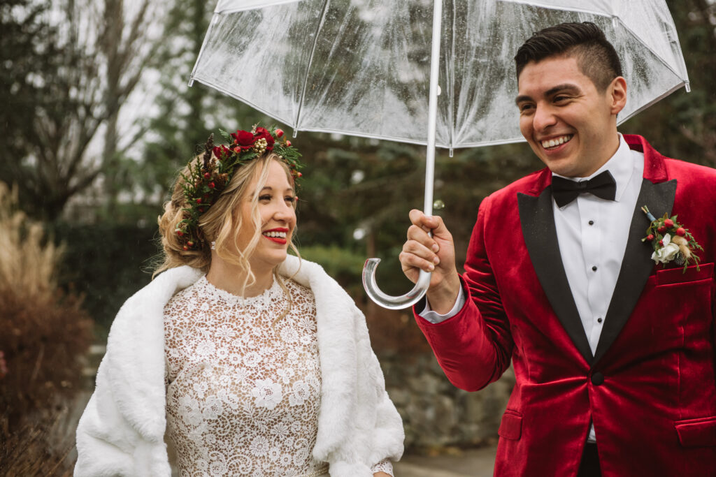 Marvin, he/him, wearing a red velvet suit jacket holds a clear umbrella over himself & Julie, she/her, who is wearing a white lace dress with a white capelet and a red floral headband. 