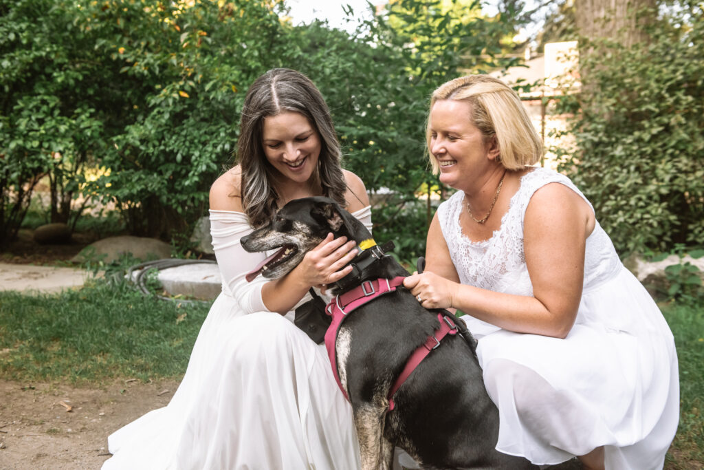 Two marriers in white dresses kneeling beside their rescue mixed breed medium dog who is sat in between them. They are both smiling, looking at their dog.