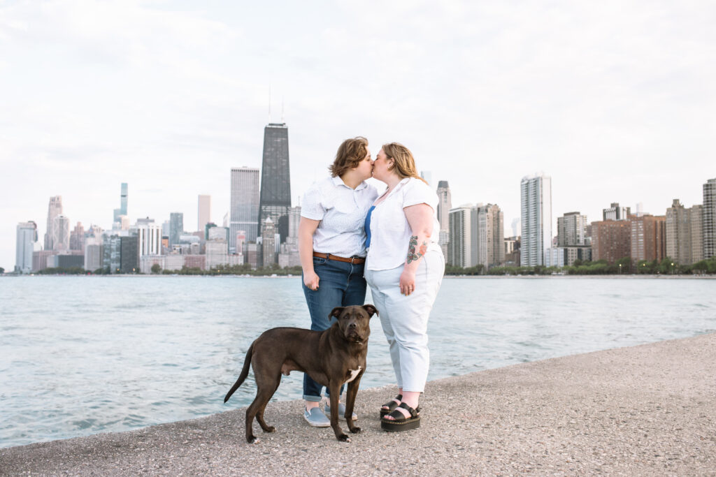 An engaged couple kissing with their rescue pitbull mixed breed medium dog standing in front of them. The dog is looking straight to the camera. In the background is the Chicago skyline at North Avenue Pier.