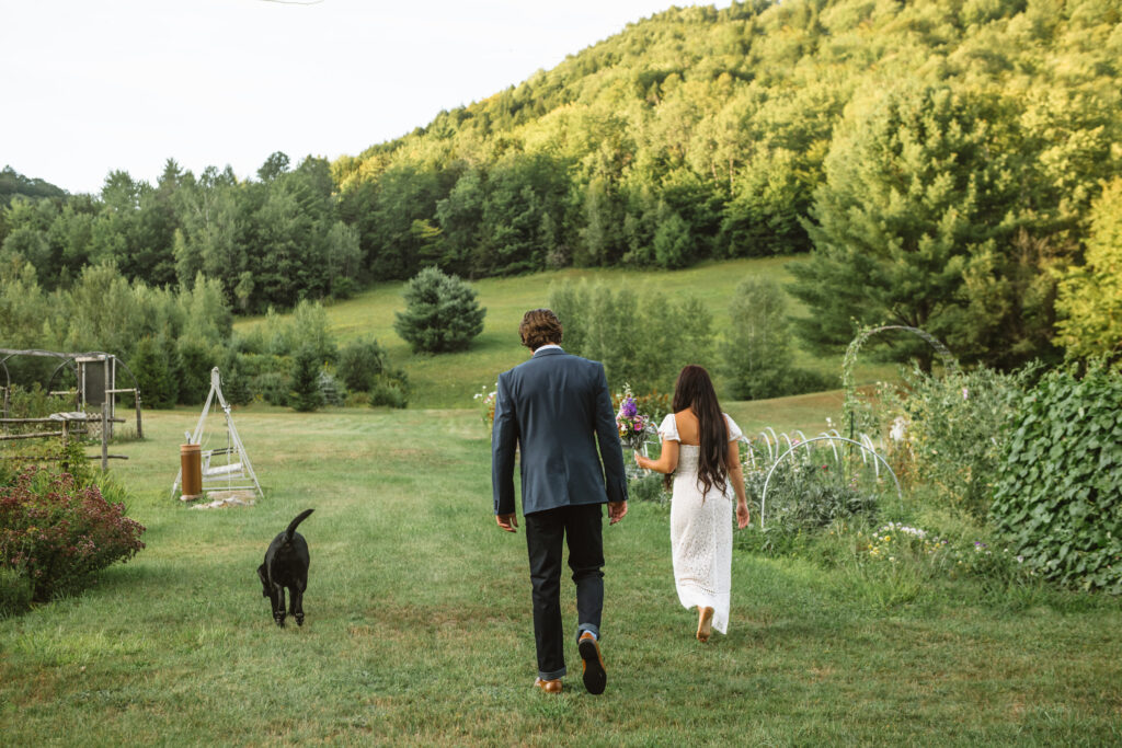 Two marriers walking away from the camera walking alongside their black lab. There are mountains and trees int he background and they are walking down an aisle in a home garden. She is walking barefoot and holding a bouquet while he is wearing a suit and brown dress shoes.