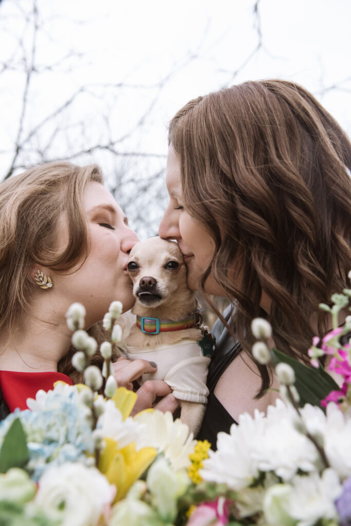 Two marriers each kissing their rescue chihuahua small dog on either side of her head, their eyes closed. The dog is looking straight to camera and her tongue is slightly poking out of her mouth. There are both of their bouquets in the bottom portion of the frame, framing the couple and dog.