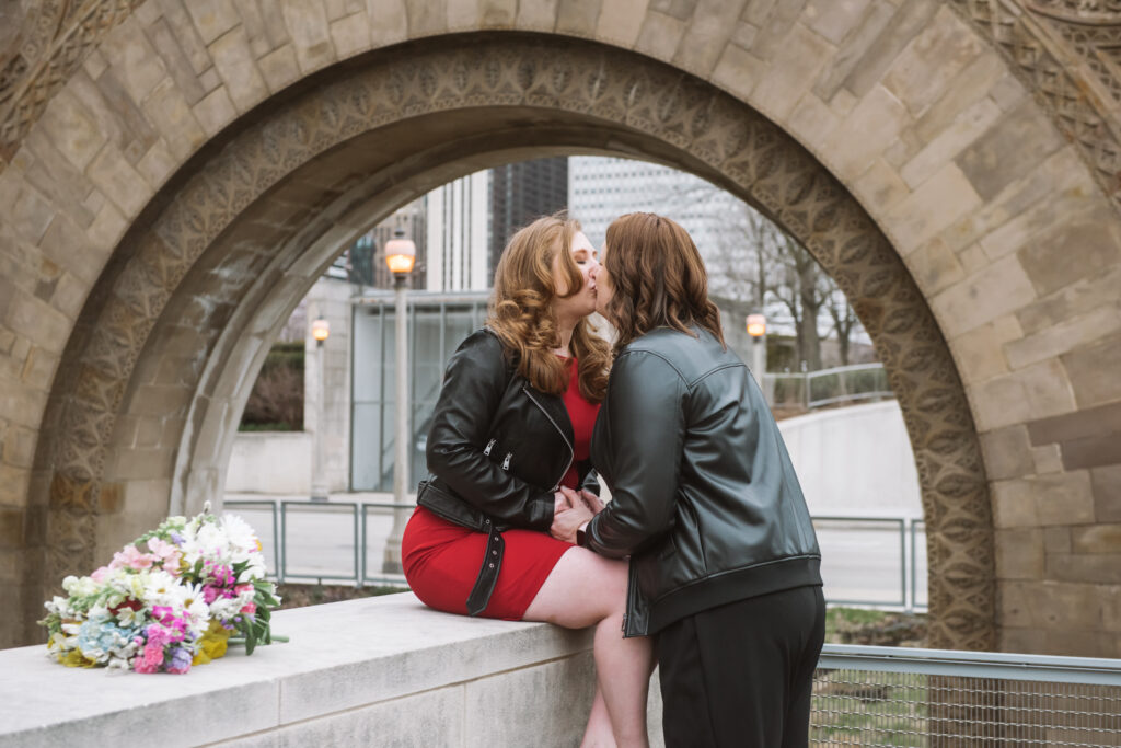 Britt, she/her, & Rya, she/her, kiss on a ledge outside the Art Institute of Chicago with their floral bouquets laying next to them.