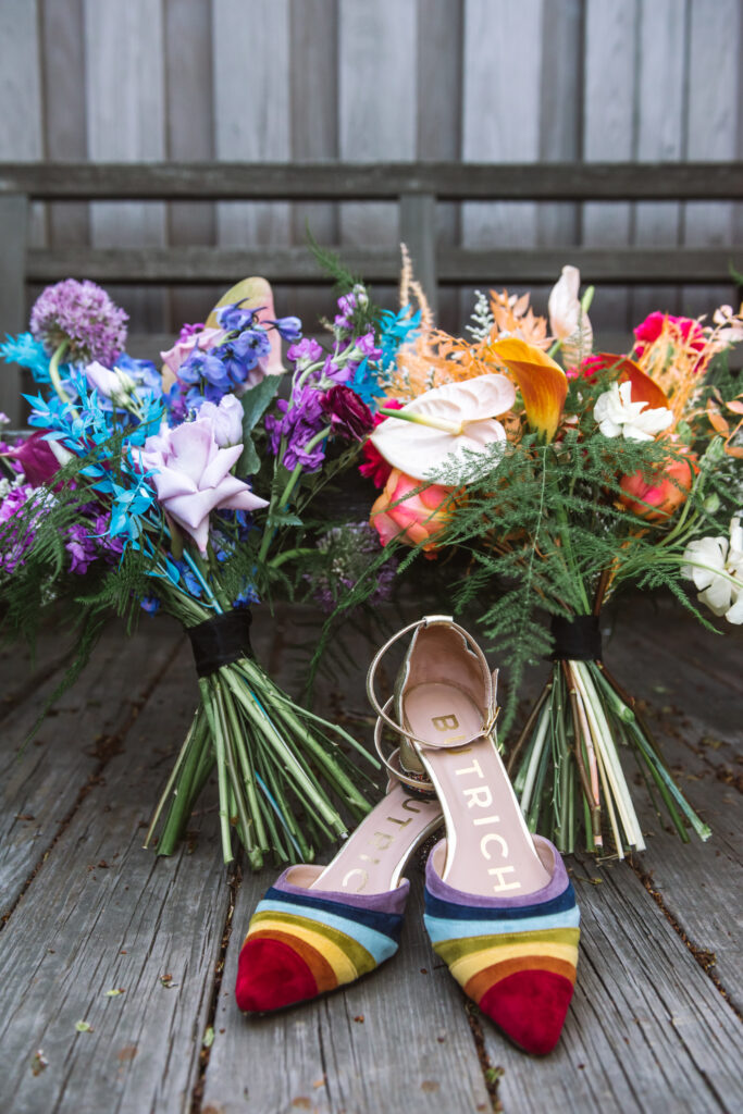 A pair of rainbow striped kitten heels sit in front of a blue & purple bouquet and an orange & pink bouquet.