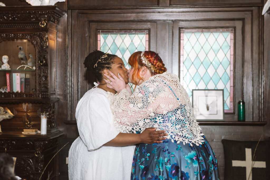 Aja, she/her, & Kyra, she/her, kiss in front of a stained glass window.
