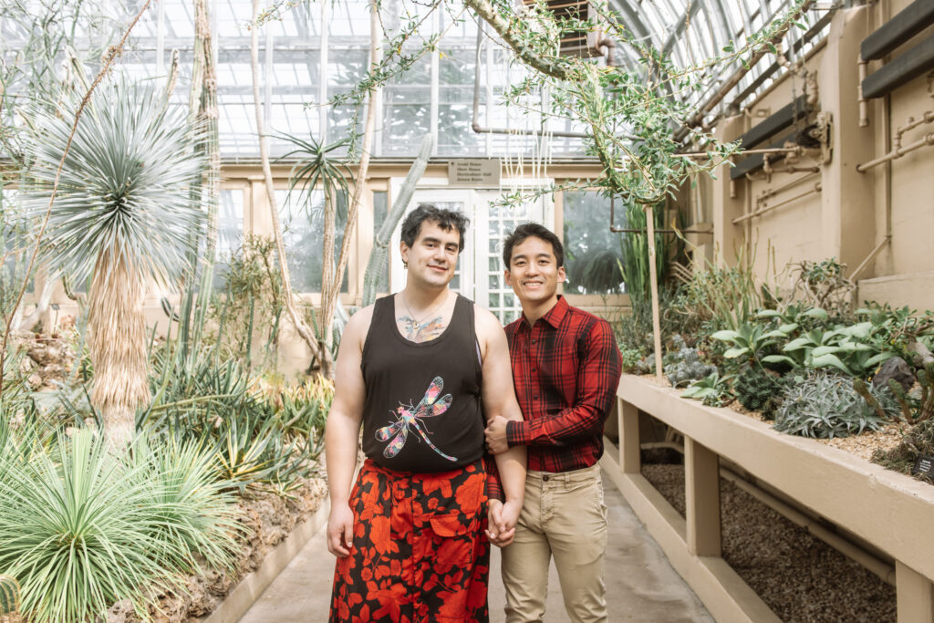 Couple standing in the desert room at the Garfield Park Conservatory in Chicago, IL. They are standing side by side with their hands interlocked. They are facing and looking directly to the camera, smiling.