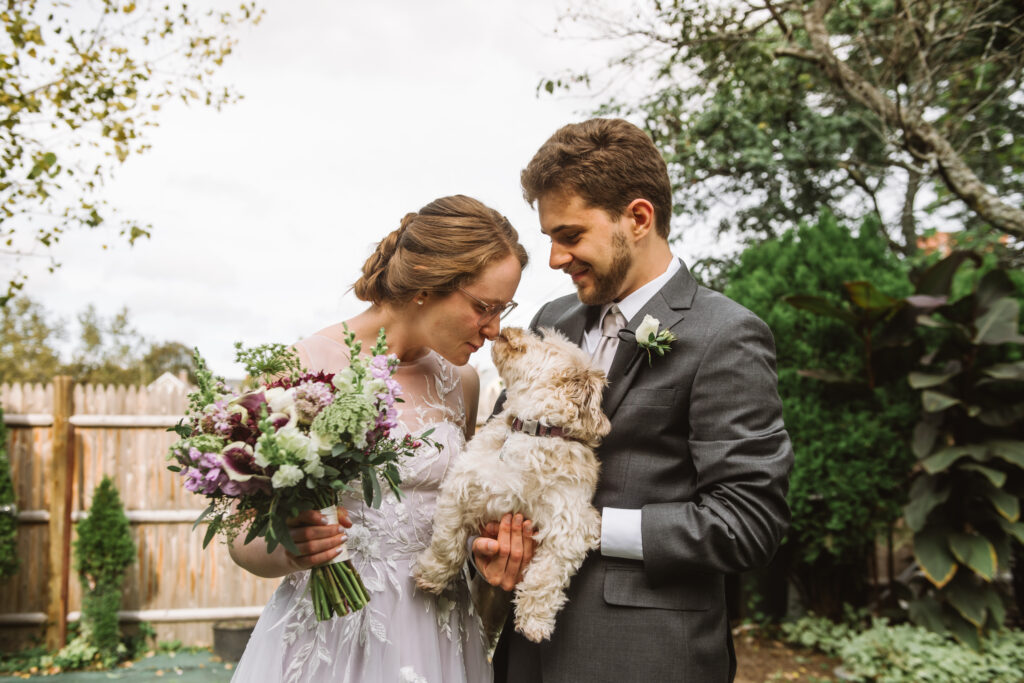 Jeff (he/him), in a grey suit,  holds small tan dog, Princess, whilst standing next to Erin (she/her), in a white bridal gown, kisses Princess & holds her purple & green bouquet. 