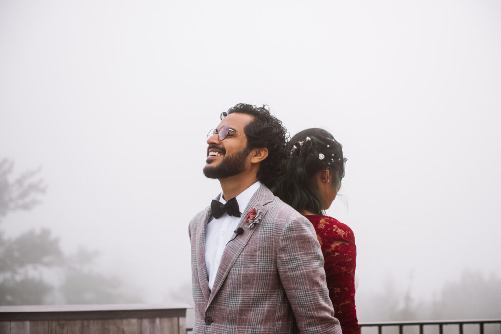 Bride and groom standing back to back for a hand holding ceremony before the actual ceremony. The focus is on the groom whose face is visible. His eyes are closed and he is open-mouth smiling. It is foggy in the background.