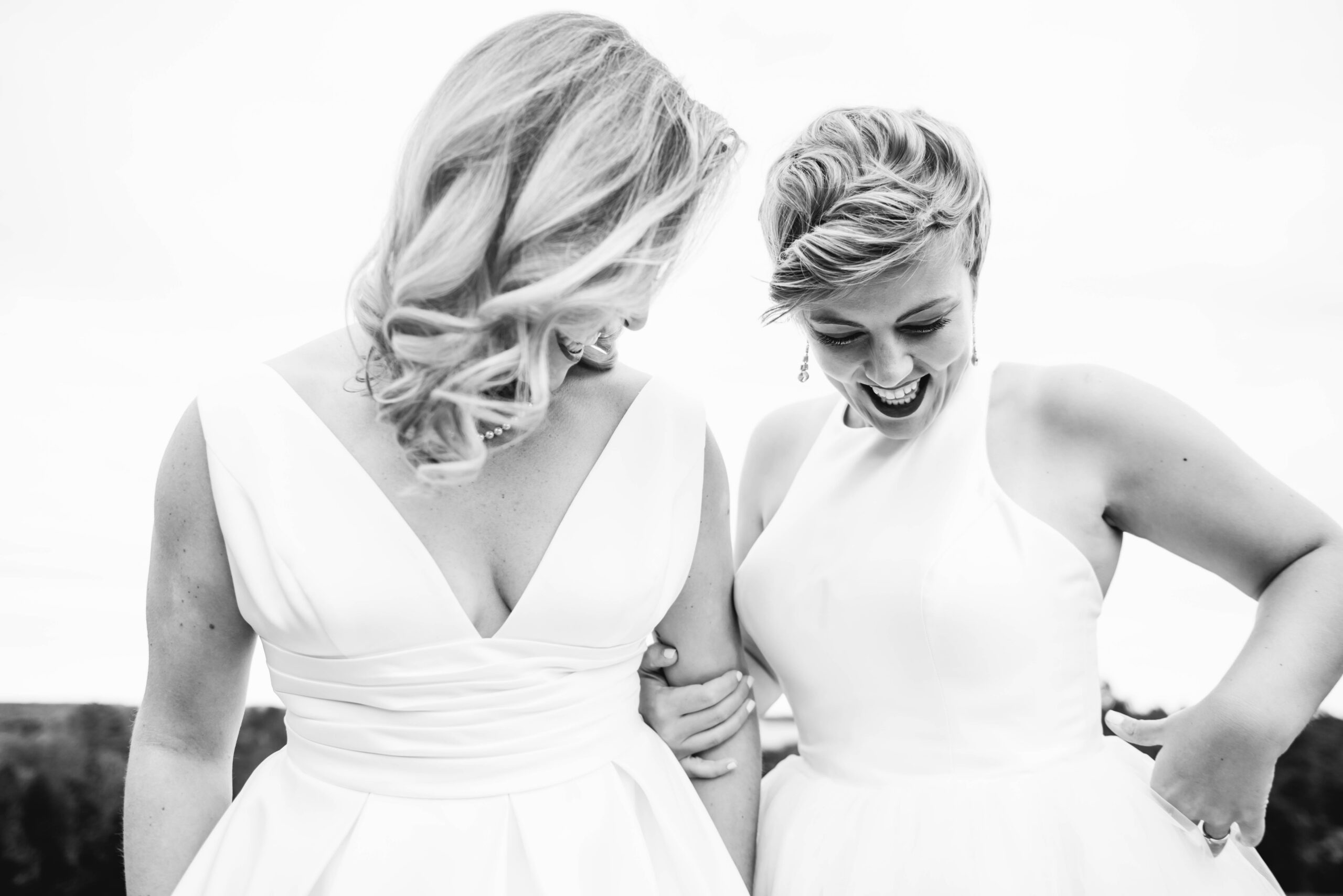 Black and white photo of two brides' torsos. They are standing close arm in arm and looking down, smiling.