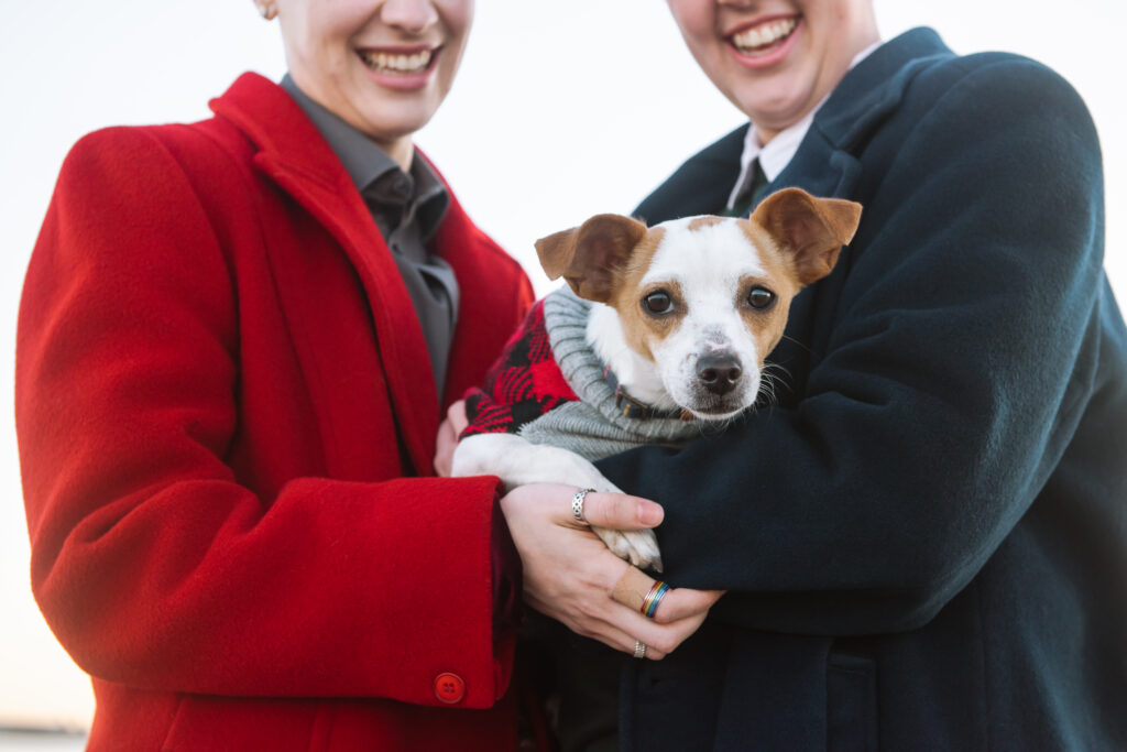 Chihuahua mixed breed looking straight to camera while being held by his two nonbinary dads. He is wearing a sweater and they are both wearing vintage 50s wool coats.