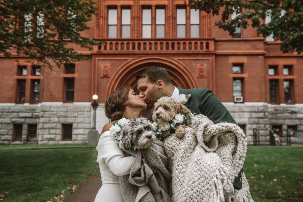 Couple kissing in wedding attire. They are each holding one of their cavapoos in a color-coordinated blanket.