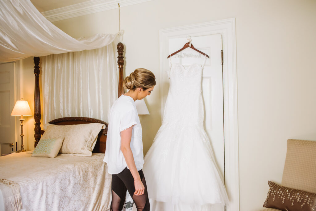 A bride looks at her wedding dress as it hangs on a door frame. 