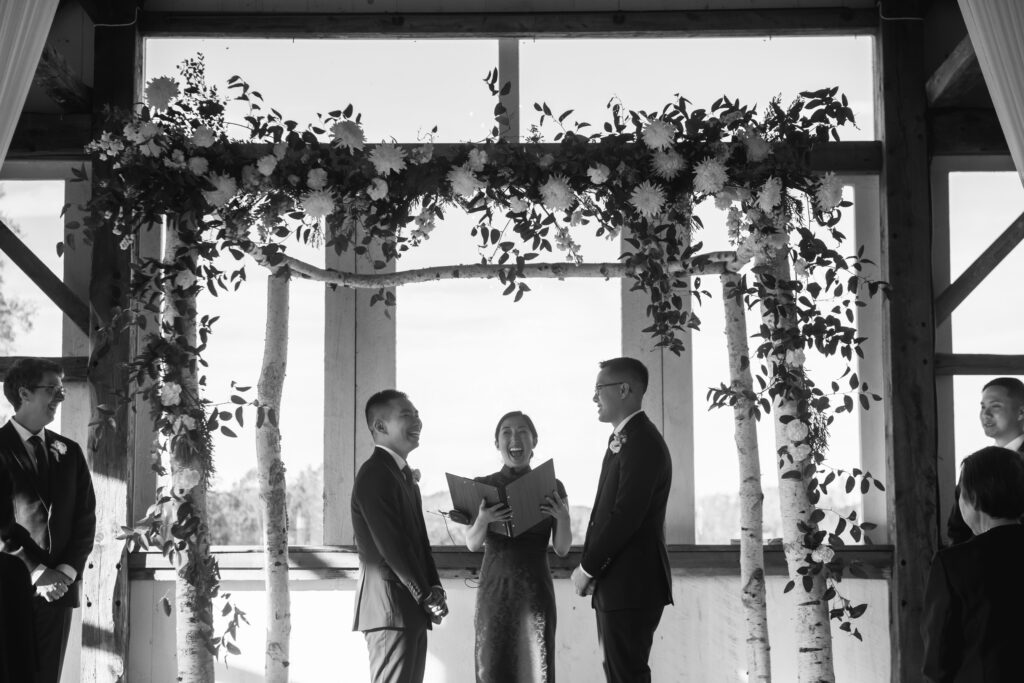 A wedding officiant laughs whilst officiating a wedding under an arch. 