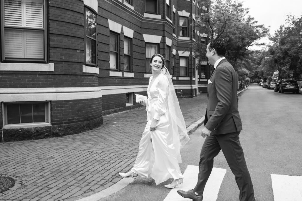 A bride and groom cross a crosswalk with a tree lined street in the background. 