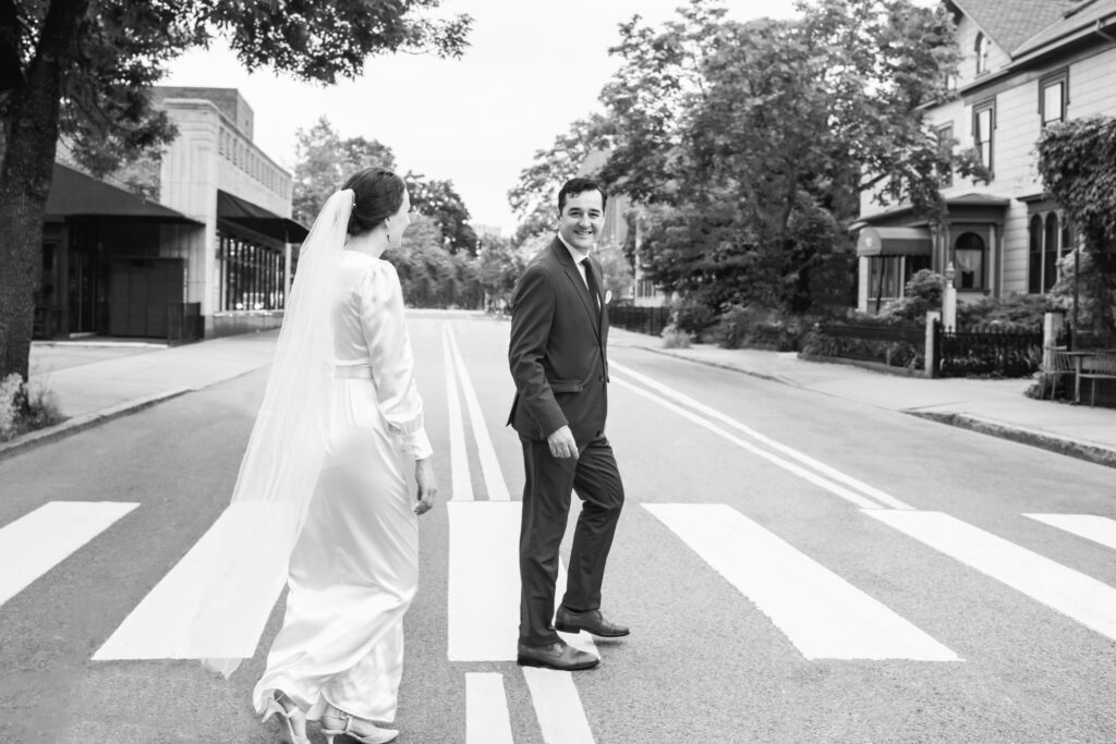 Black and white photo of couple walking on a crosswalk in the middle of the road. The groom is looking toward the camera while walking forward. The bride has her back to the camera walking towards the groom.