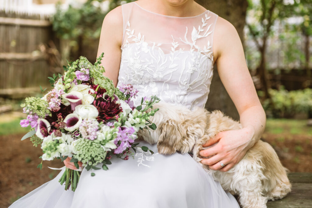 Bride's torso, sitting down, holding a bouquet. She has her dog sitting next to her on the bench and her dog's face is half hidden by the bouquet.