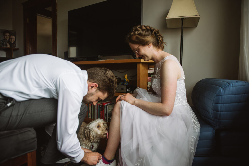 Groom tying his bride's shoes. They are both sitting down in wedding attire. Their mixed breed dog is in between them on the floor, licking the bride's shin.