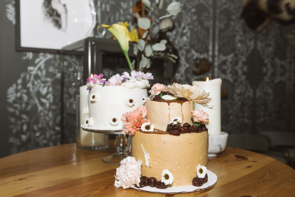 Two cakes, one is a two-tiered light brown cake and the other is white.  Both have fresh florals as decorations.